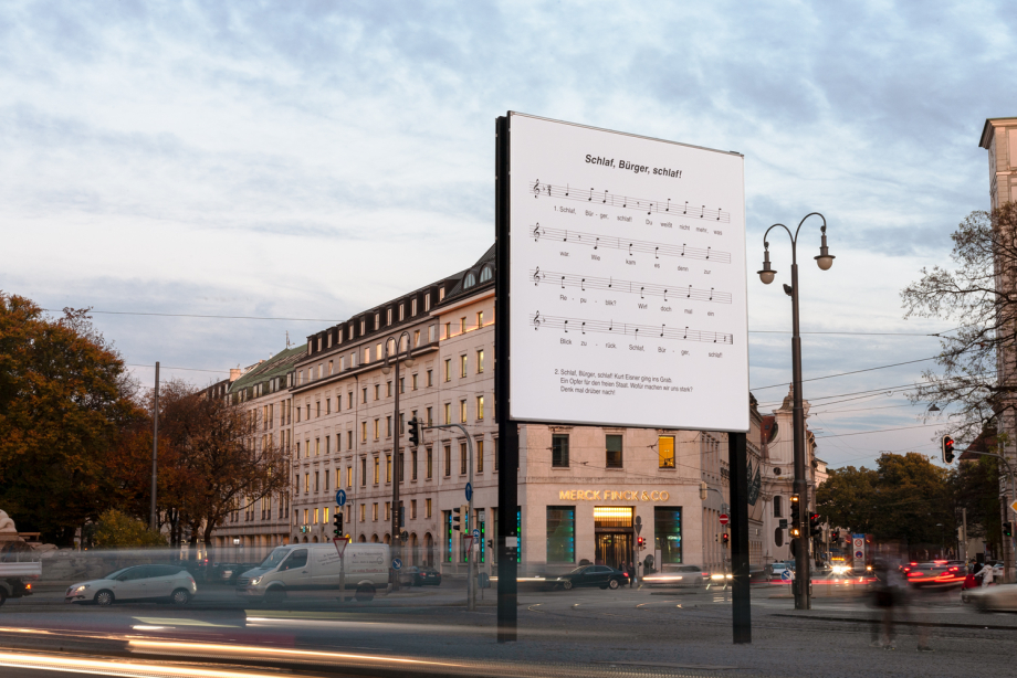 Seitliche Ansicht des Billboards am Lenbachplatz während der abendlichen Rushhour. In schwarzer Schrift auf weißem Grund erscheint der Notensatz des Kinderliedes "Schlaf, Kindlein, schlaf!" mit abgeändertem Text zu "Schlaf, Bürger, schlaf!".
